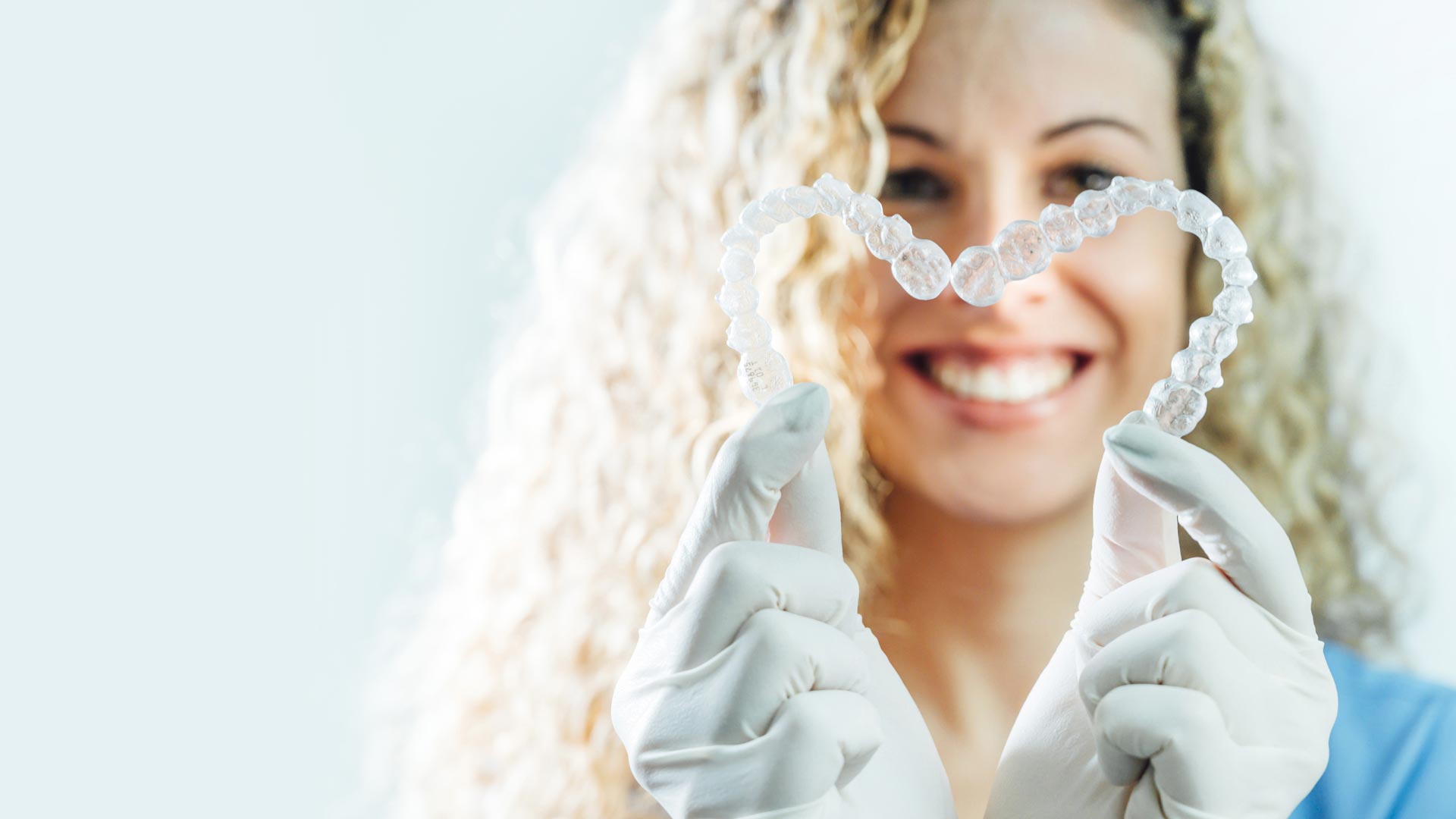 A dentist smiles and holds up two clear aligners in the shape of a heart.